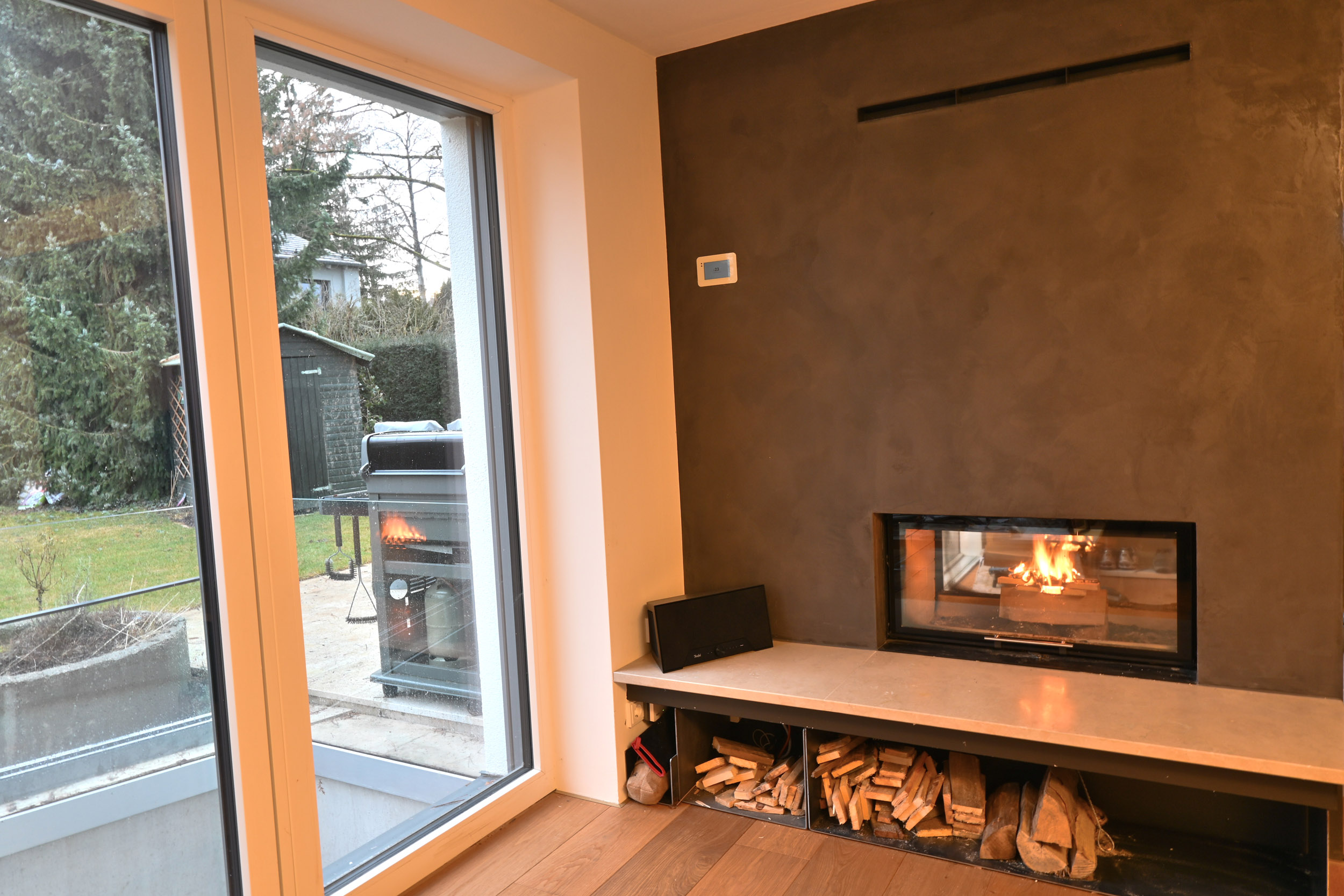 brennender Kamin in einer dunklen Wand verbaut als Raumteiler mit einer Sitzbank davor